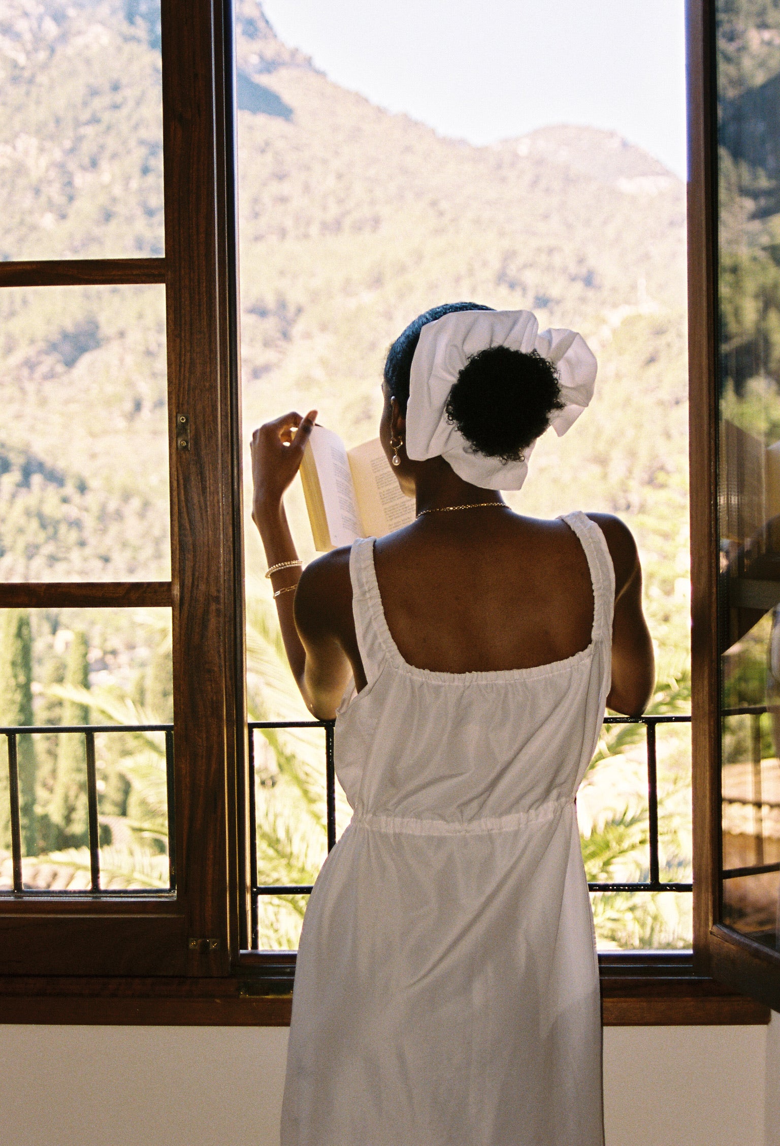 Woman in a white dress standing by a large window with a mountain view wearing beautiful gold jewerly handmade by me 