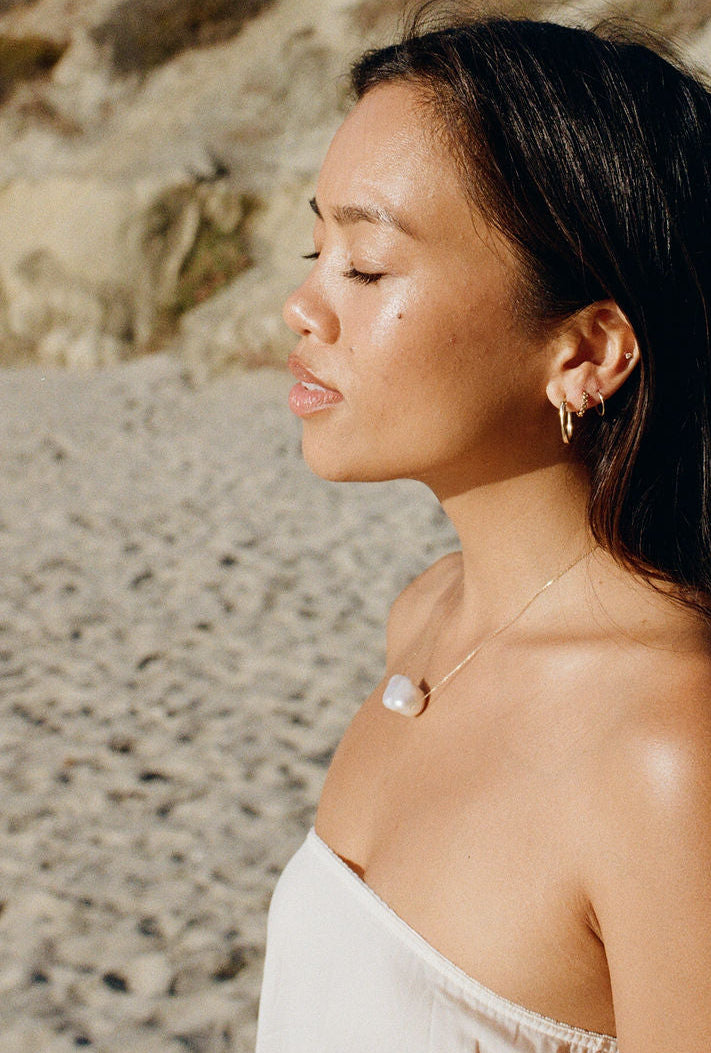 A woman with dark hair stands in profile on a sandy beach with rocky cliffs behind her, wearing a light beige strapless top and small hoop earrings.